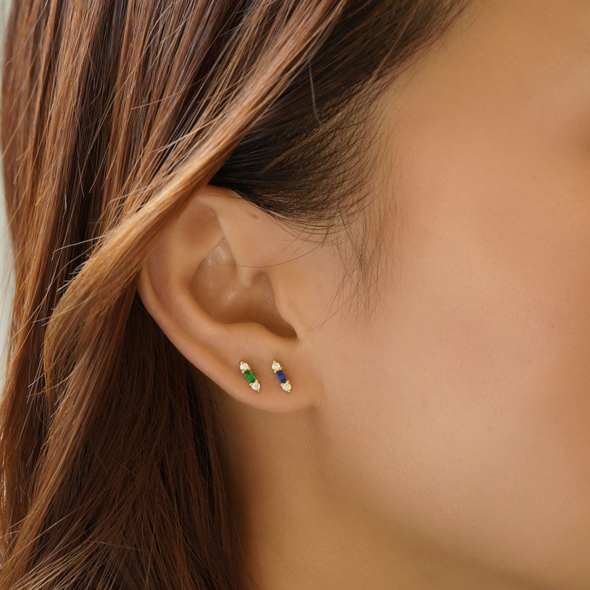 Gold vermeil tiny blue baguette stud shown beside a green baguette stud on a model's lobe