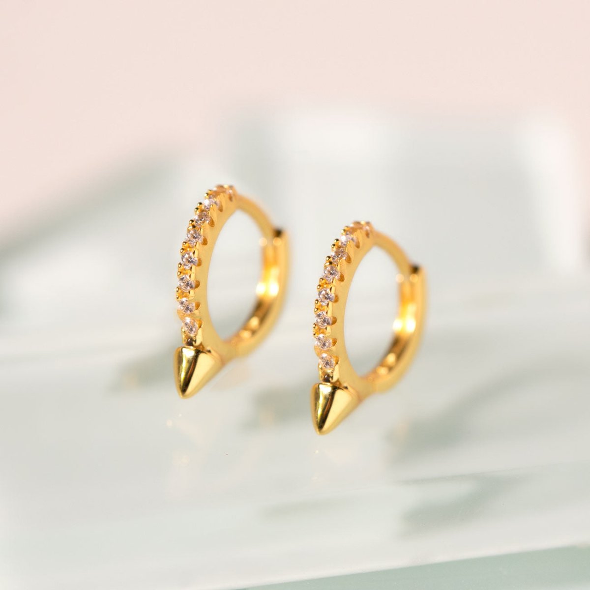 Gold vermeil pavé spike huggie hoops with clear stones, shown on a pale prop surface
