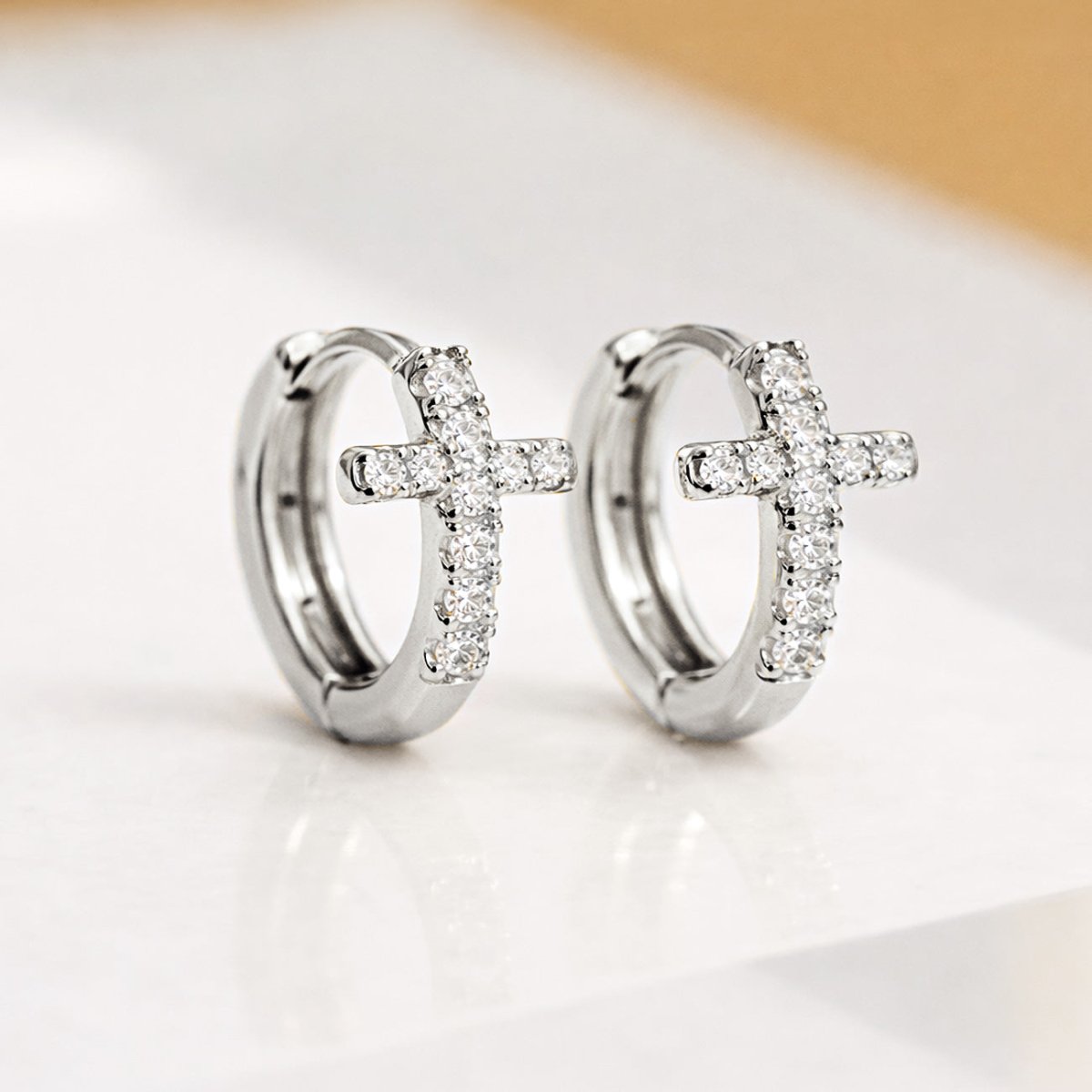Sterling silver cross huggie hoops with pavé clear crystals, shown angled on a cream surface