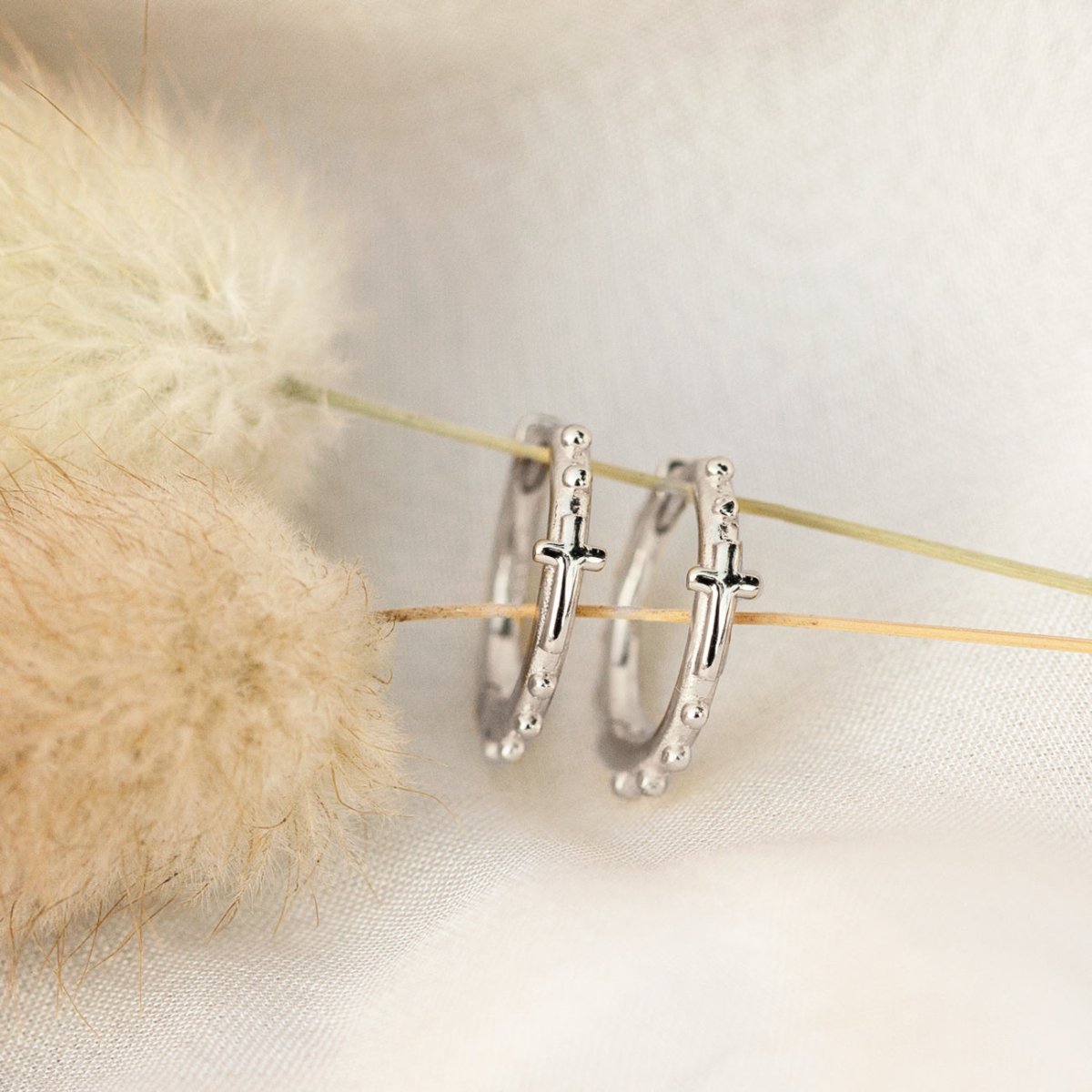 Sterling silver beaded cross huggie hoops propped against dried flowers on cream fabric