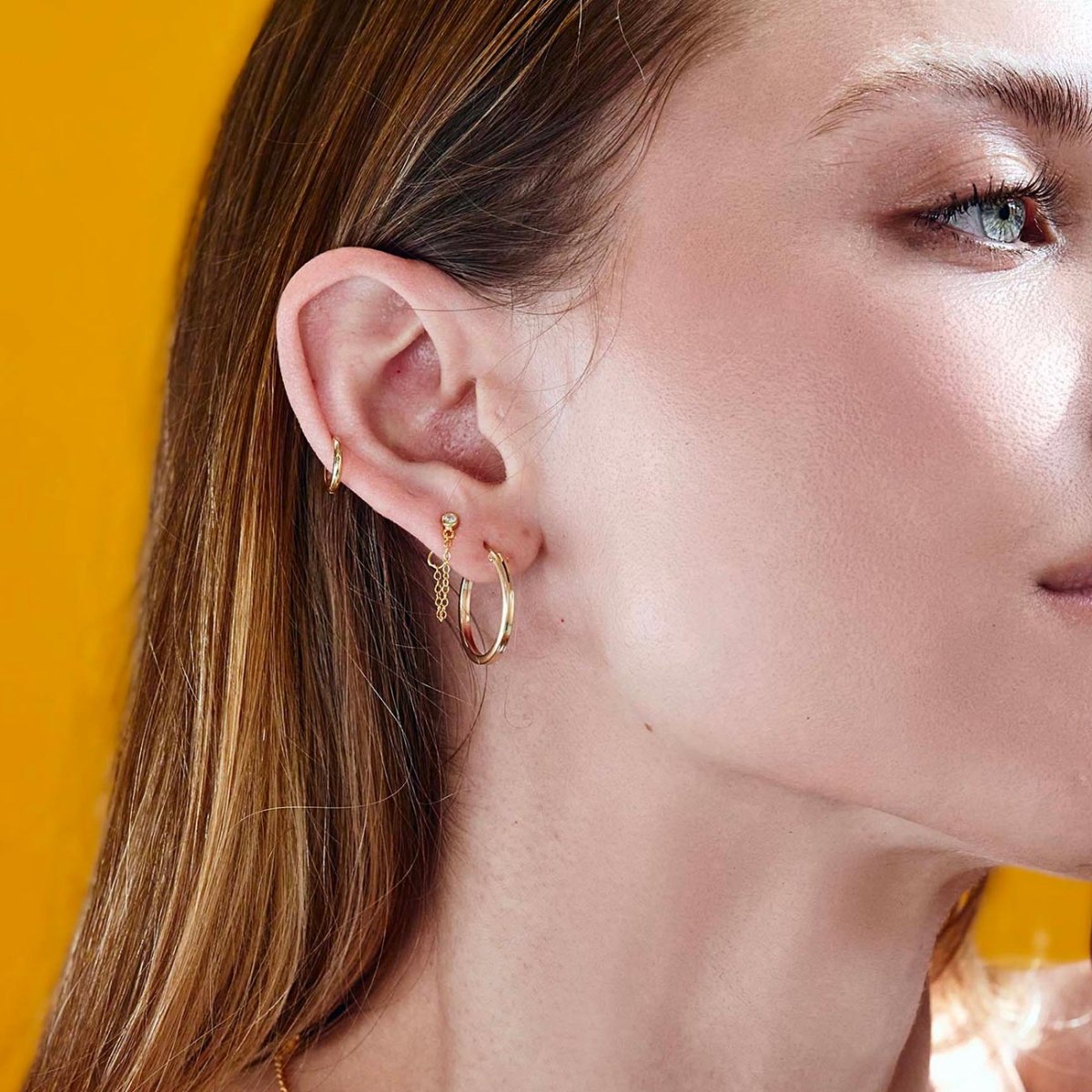 Gold-filled square-edge hoop earrings shown on a model's ear against an orange background