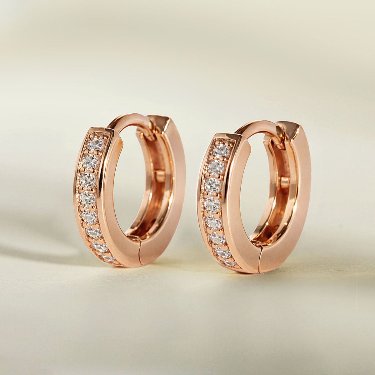 Rose gold vermeil pave huggie hoops with a row of clear white zirconia, three-quarter view on a cream background