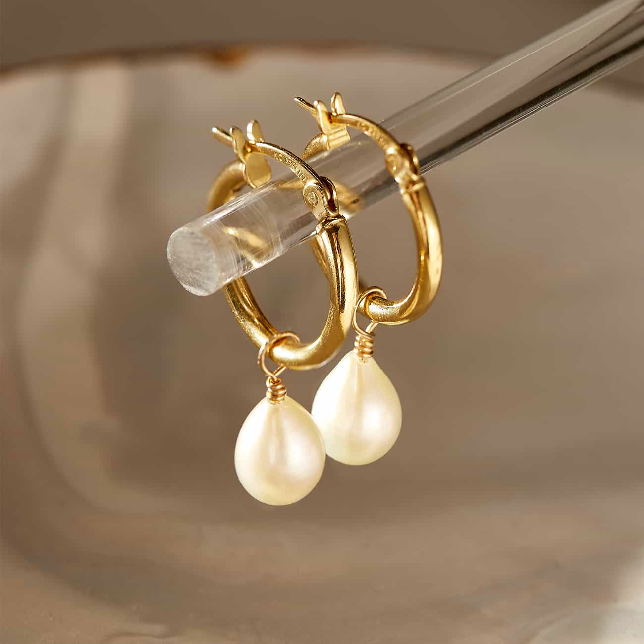 Gold-filled huggie hoops with a dangling white teardrop pearl, held on a clear rod, close-up