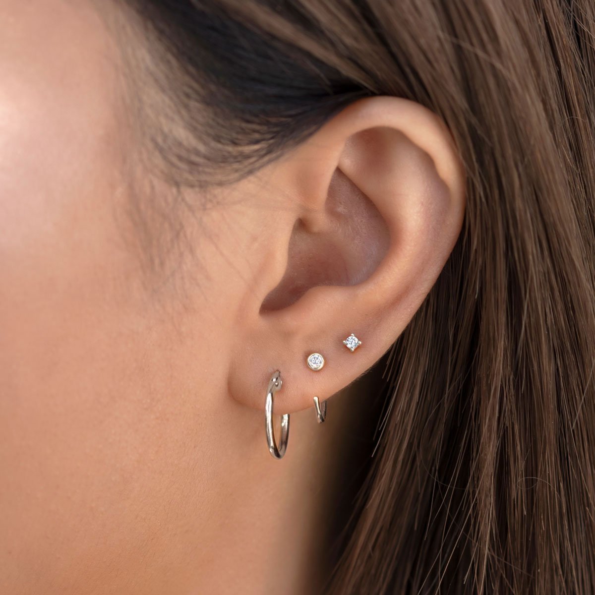 Sterling silver small hoops shown on a model's lobe alongside round and square clear-stone studs