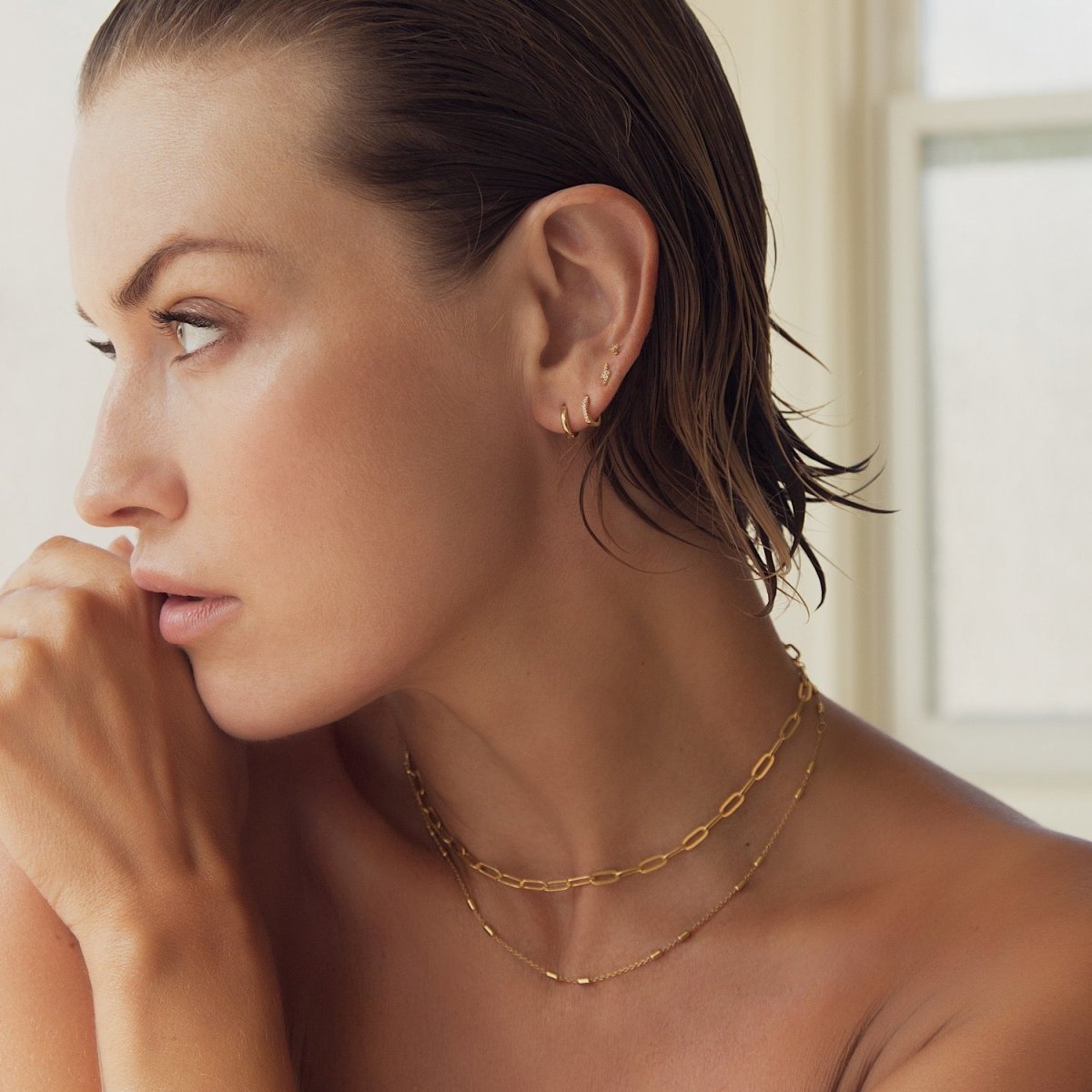 Model wearing layered 14K gold paperclip and fine chain necklaces with tiny ear studs