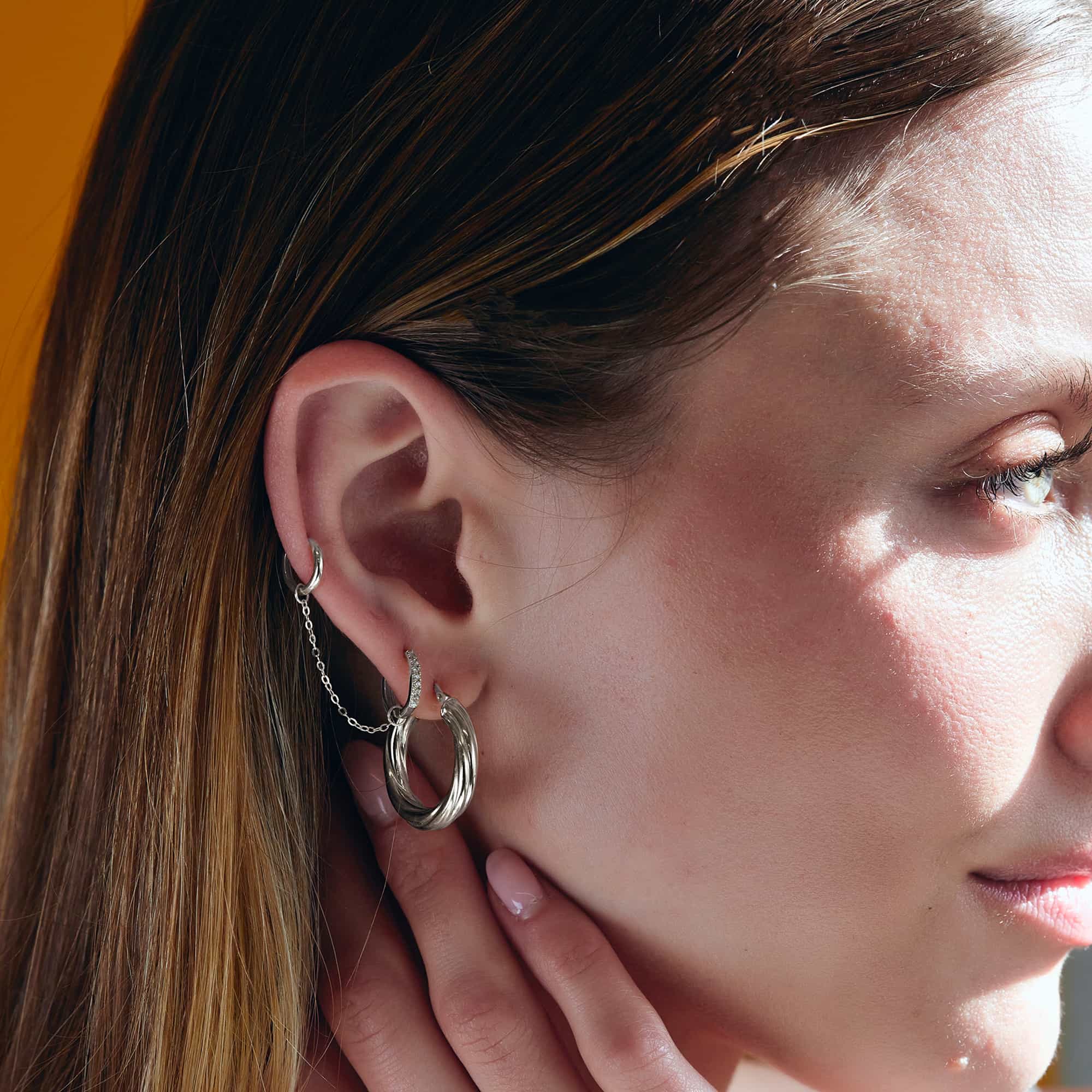 Close-up of woman wearing 14k white gold seamless clicker ring, sterling silver thick twisted hoop, 14k white gold chain ear jacket for huggies, 14k white gold single thin pave huggie, and 14k white gold mini huggie