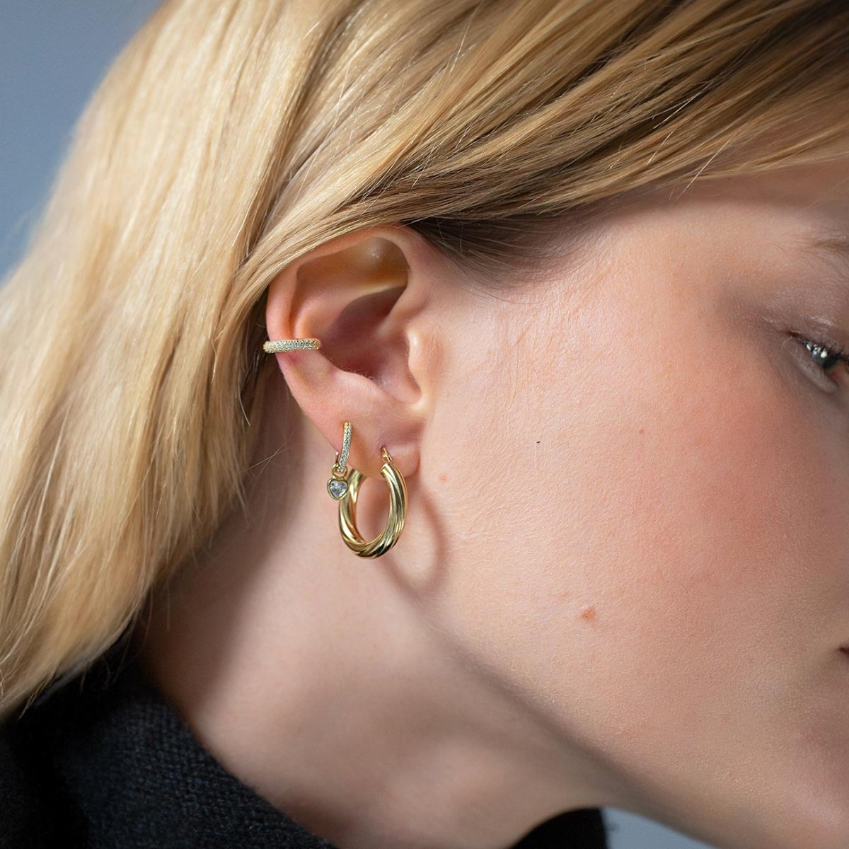 Gold twisted hoop and a pavé huggie with a clear heart charm shown on a model's ear