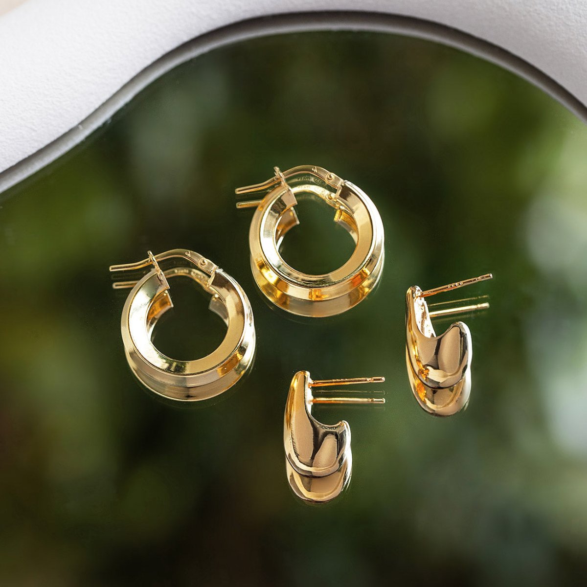 Set of gold hoop earrings on a reflective surface with a blurred green background - shop timeless everyday hoops.