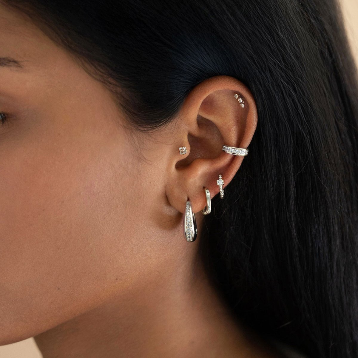 Silver hoops, ear cuffs and studs stacked along a model's ear, close-up side view