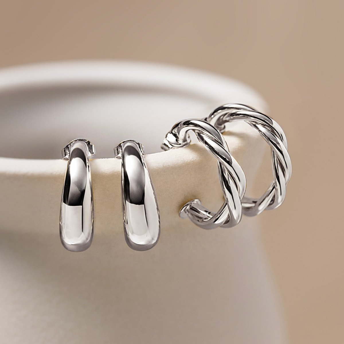 Sterling silver tapered C huggie hoops beside twisted silver hoops, on a cream background
