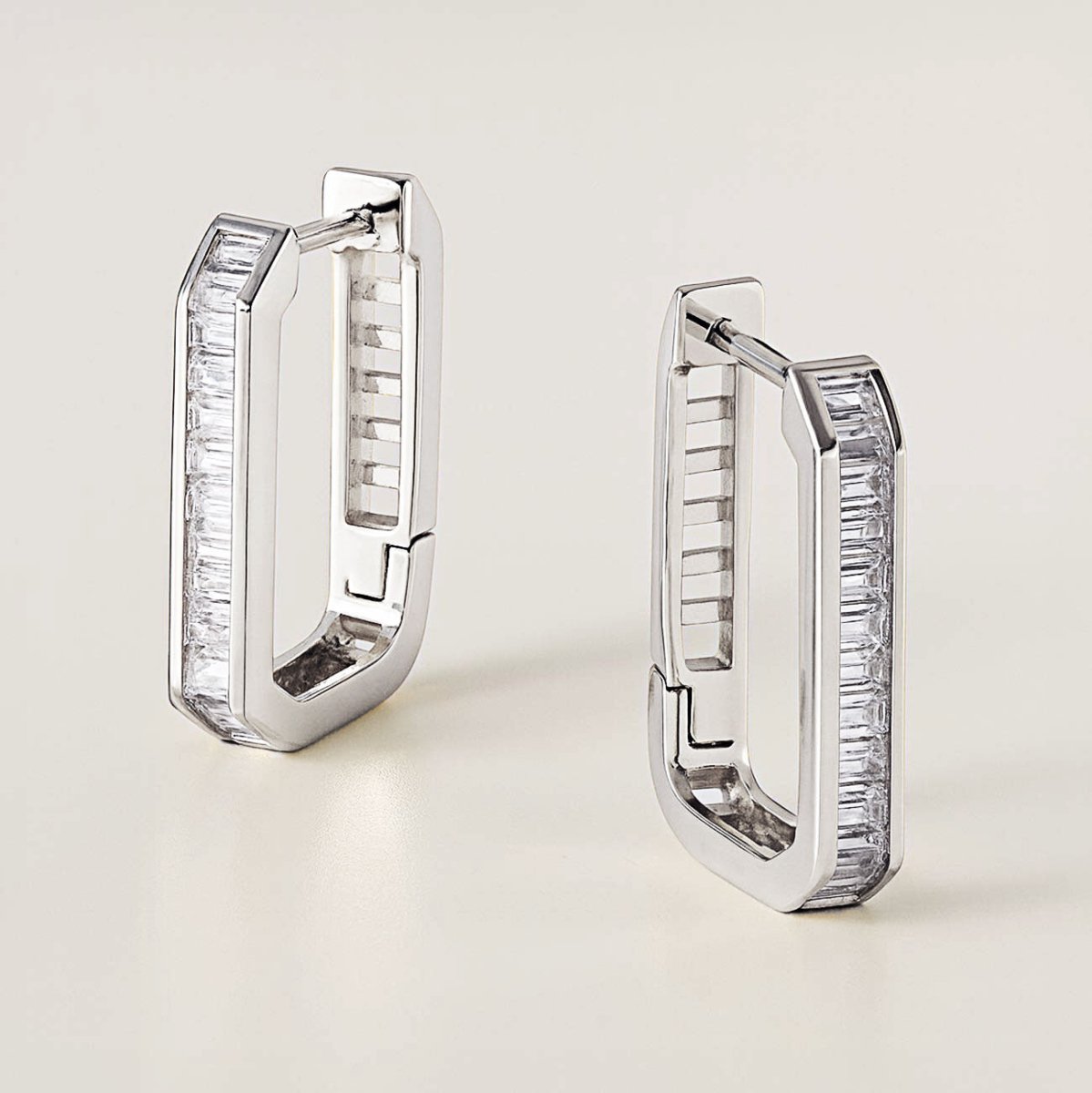 Sterling silver channel-set baguette oval huggie hoops with white zirconia, pair shown standing on a cream background