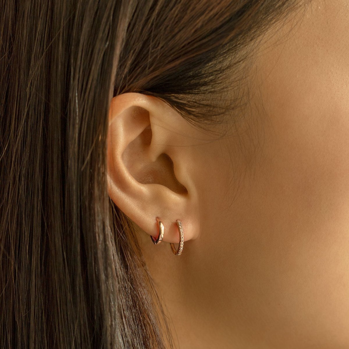 14K rose gold diamond and plain huggie hoops layered on a model's lobe