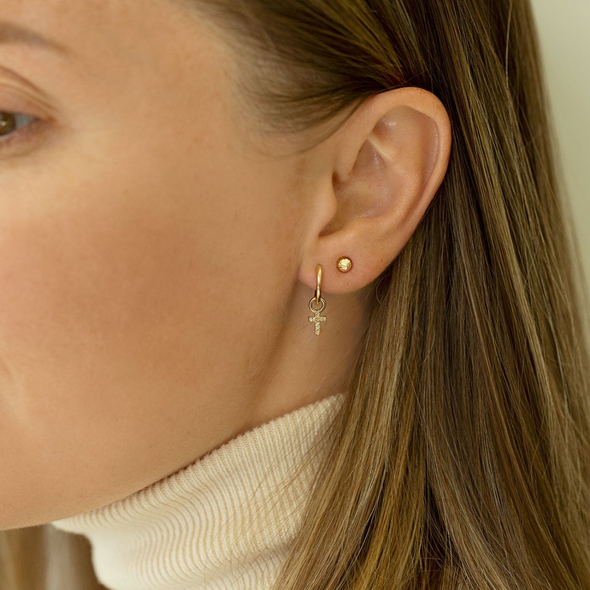 14K gold huggie hoop with a dangling diamond cross charm shown on a model's lobe