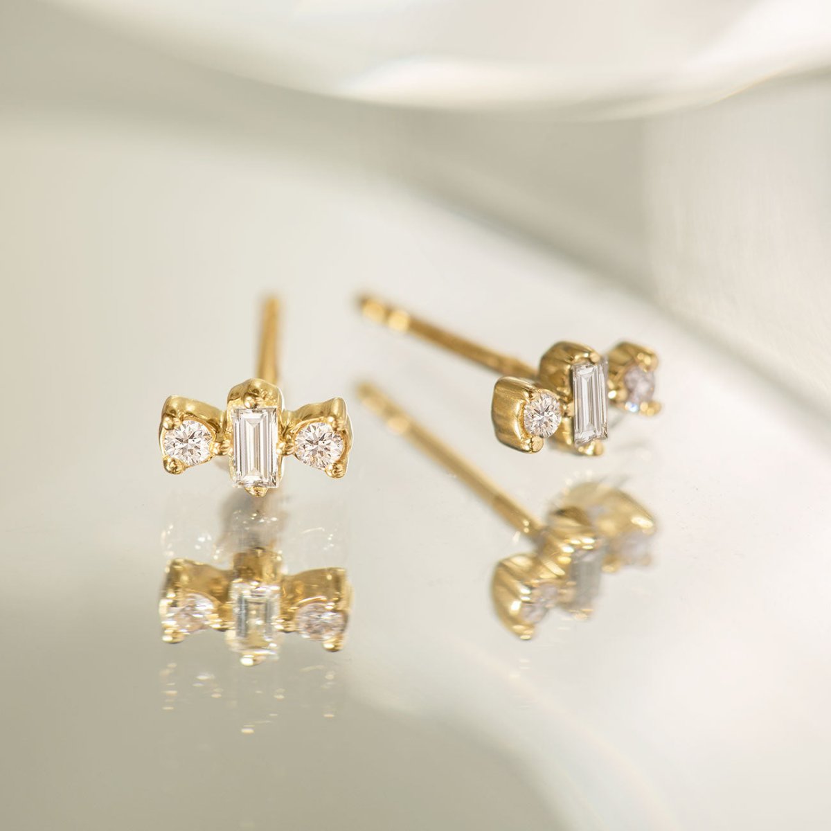 14K gold diamond baguette triad studs arranged on a cream surface, close-up detail