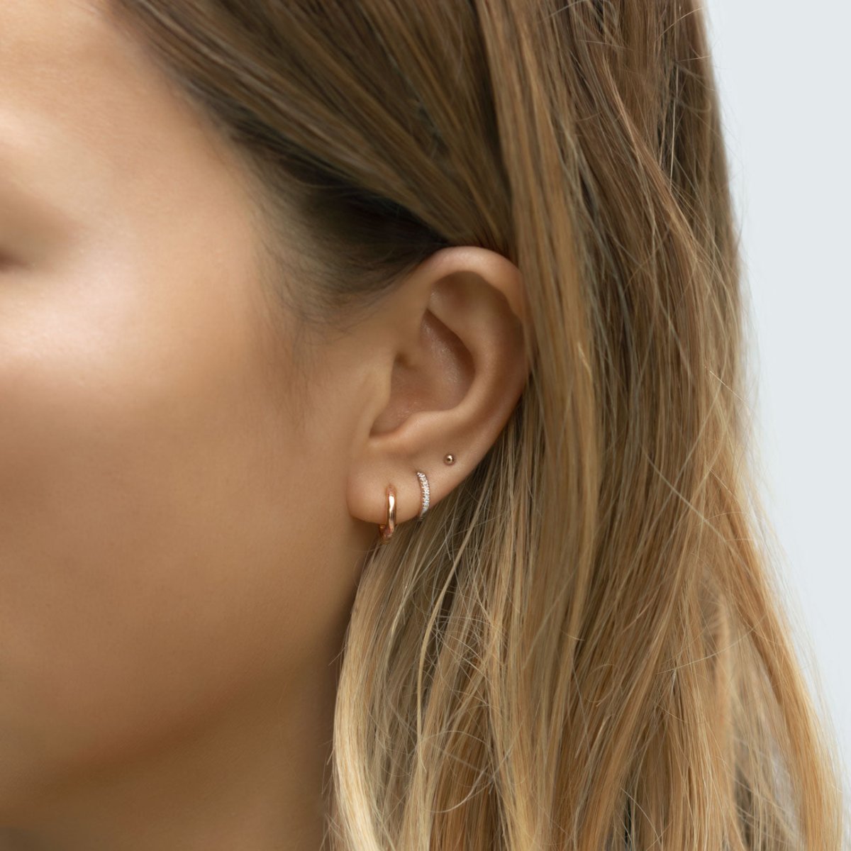 14K rose gold thin diamond huggie hoop shown on a model's ear with two stud piercings