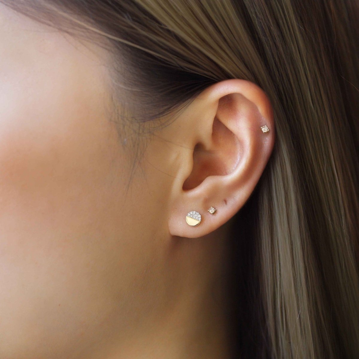 14K gold half-disc stud with pave clear stones, shown on a model's lobe with smaller studs
