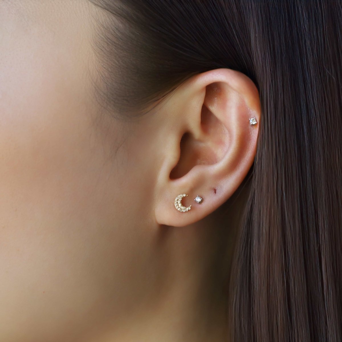 14K gold diamond crescent moon stud beside a small round diamond stud, shown on a model's lobe