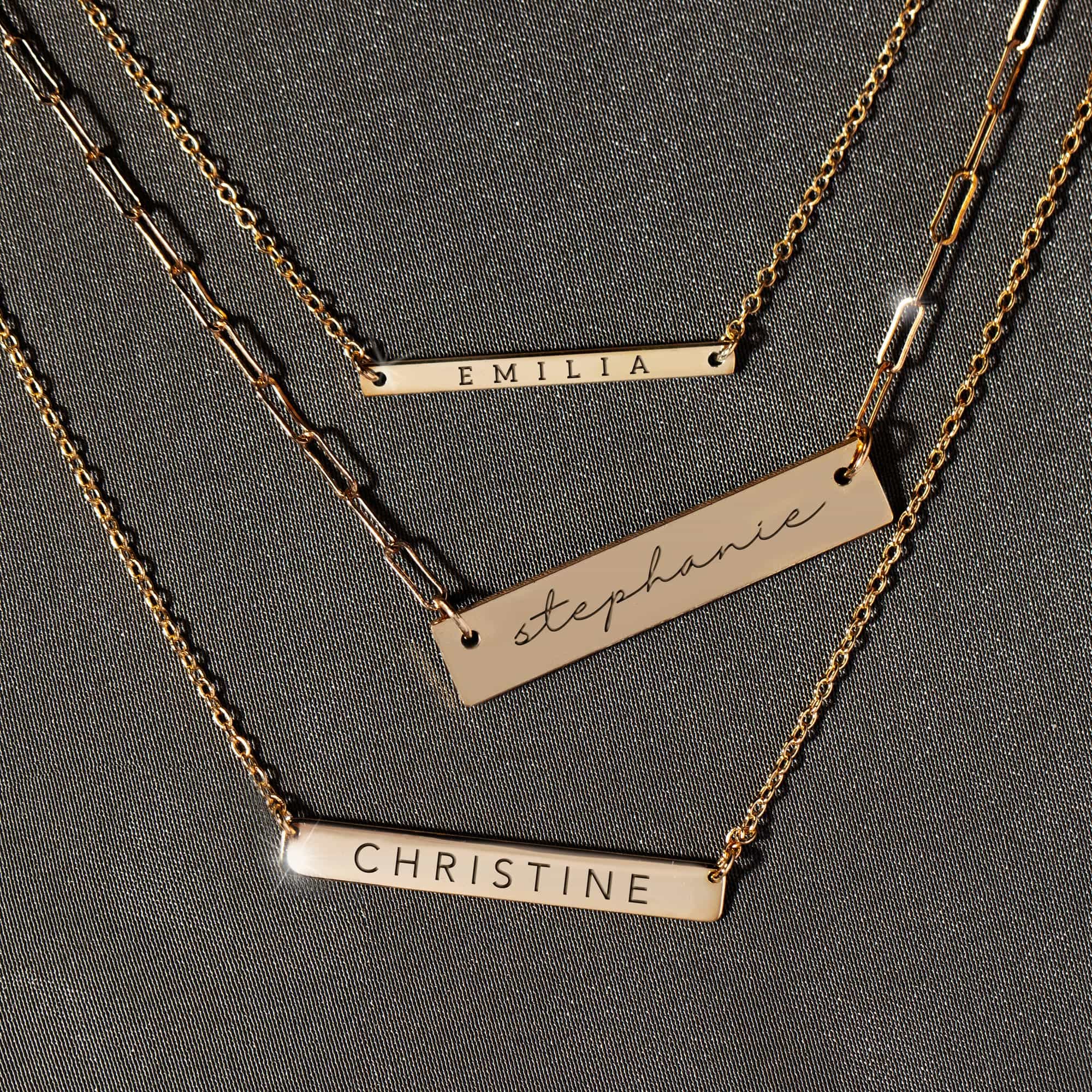 Three gold-filled engravable bar necklaces engraved with names, layered on gray fabric