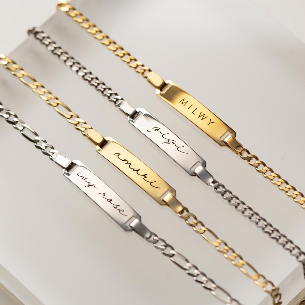 Sterling silver and gold Figaro chain ID bracelets, each engraved with a name, on a cream background