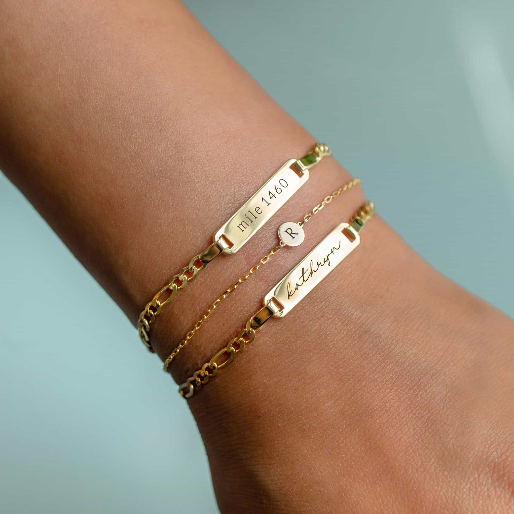 Gold ID bracelets engraved with names and an initial disc, on a model's wrist
