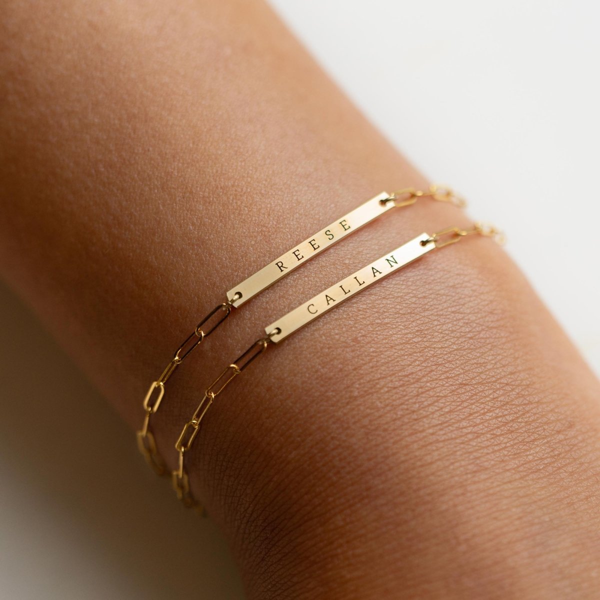 Two gold-filled thin bar paperclip chain bracelets engraved with names, shown on a model's wrist