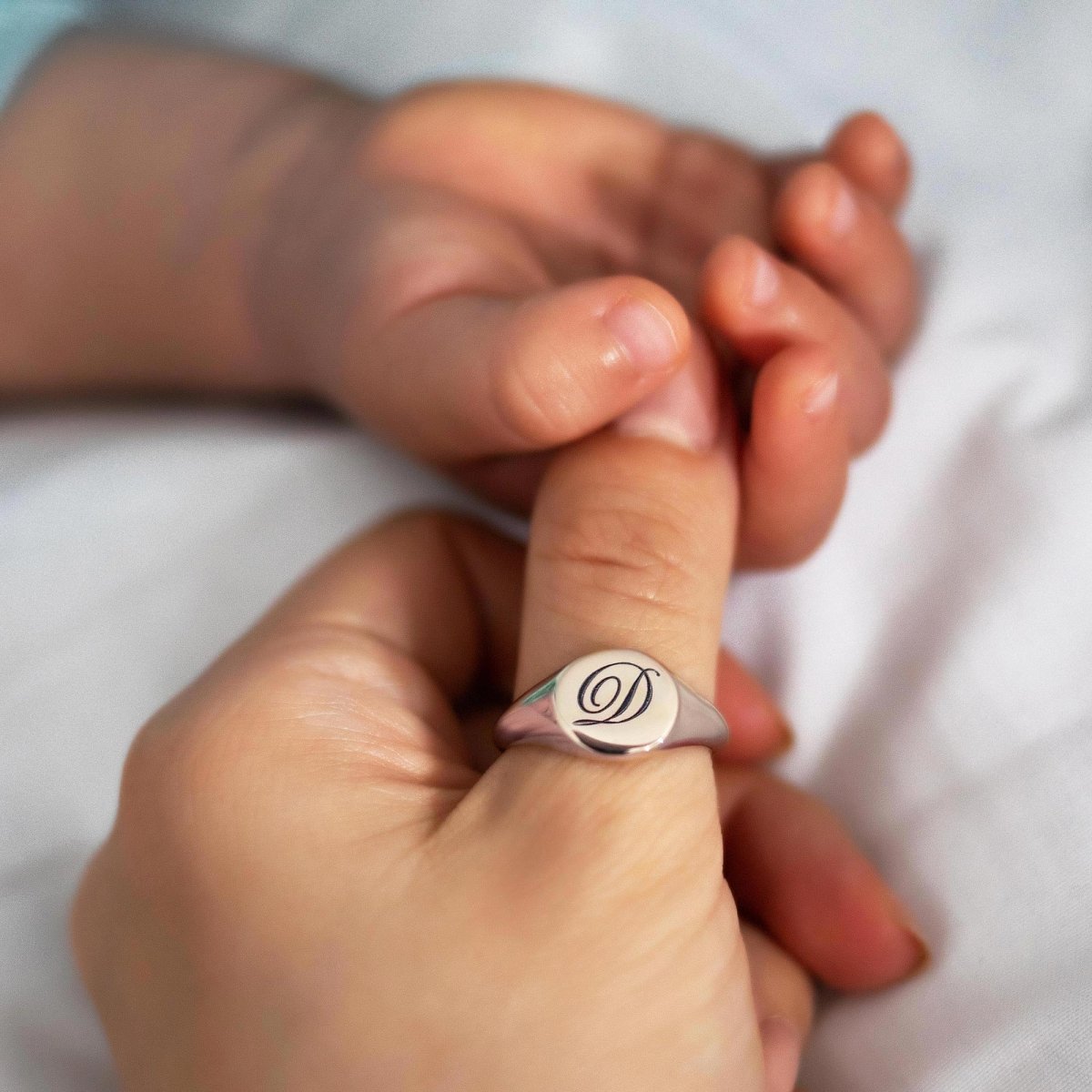Sterling silver oval signet ring engraved with an initial, shown on a hand holding a baby's hand