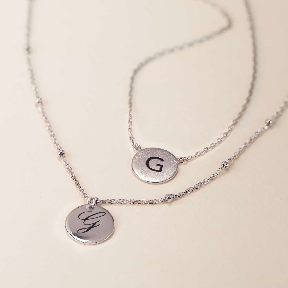 Two sterling silver initial disc necklaces, engraved with an initial in block and script, on a cream background