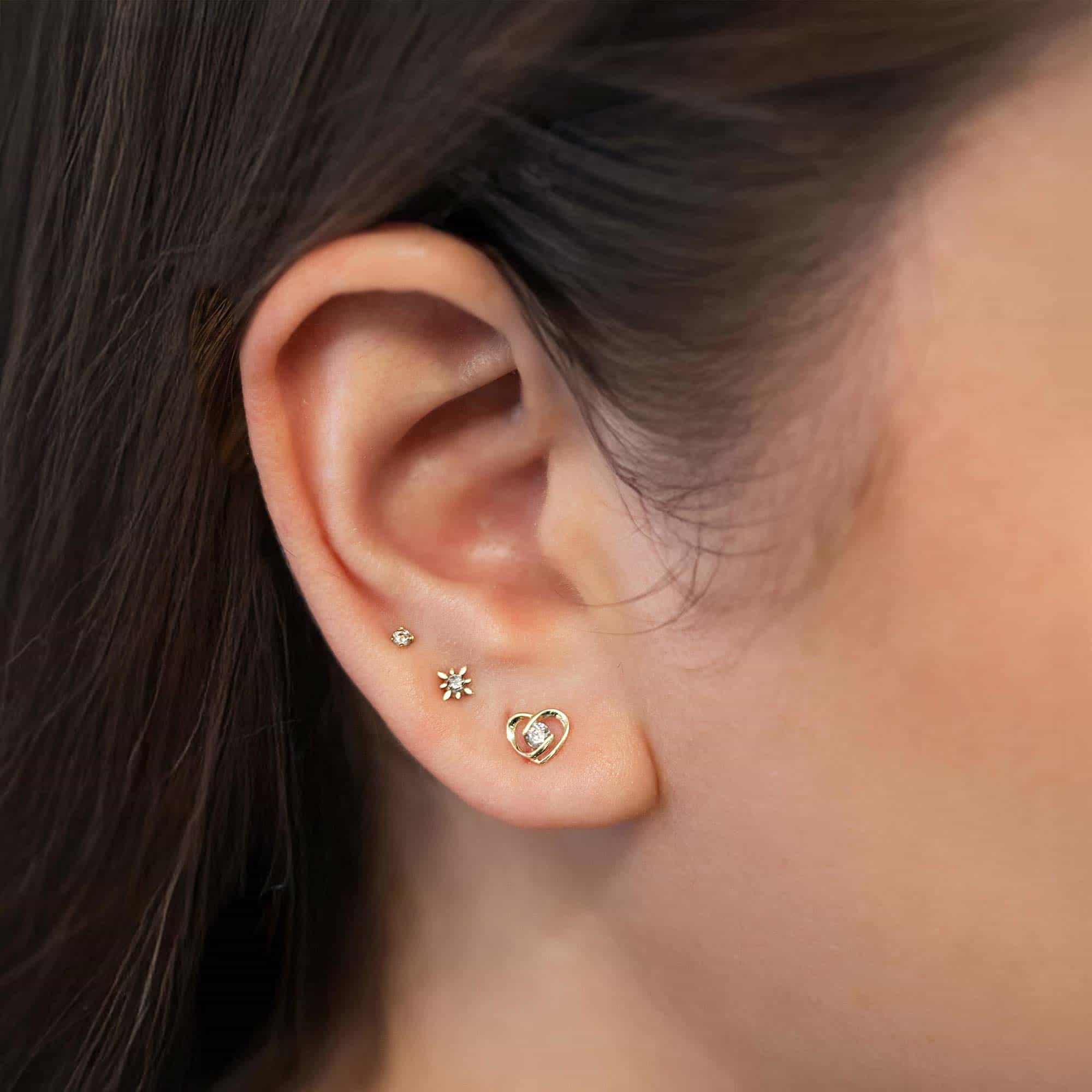 14K gold heart outline white zirconia stud shown on a model's lobe in a stack with smaller studs