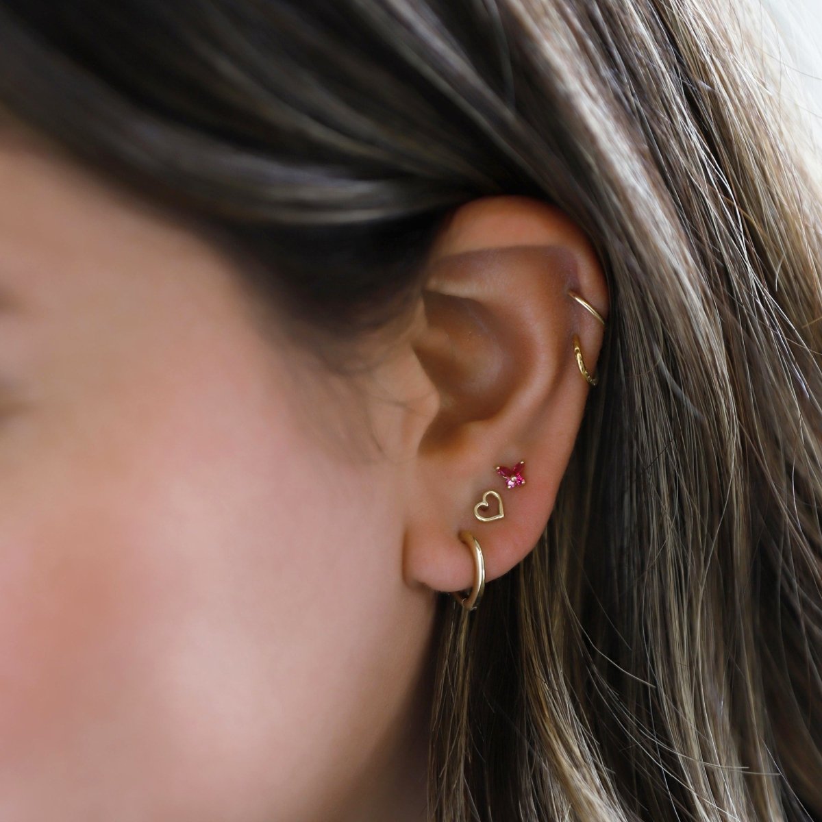 14K gold mini heart outline huggie hoop shown on a model's lobe with a pink stone stud above