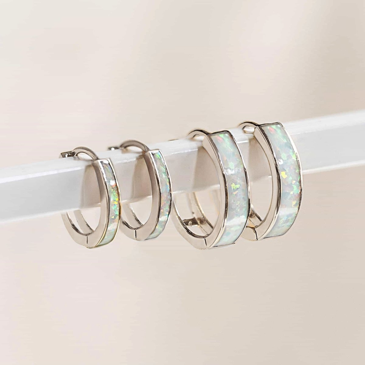 14K white gold white opal huggie hoops in two sizes, lined up on a clear acrylic bar