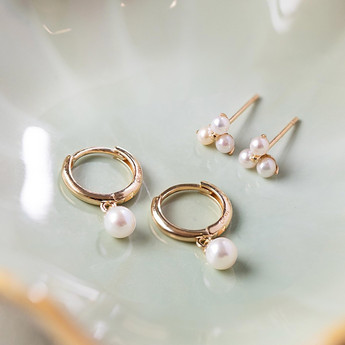 Gold pearl-drop huggie hoops and trinity pearl studs arranged on a pale green dish