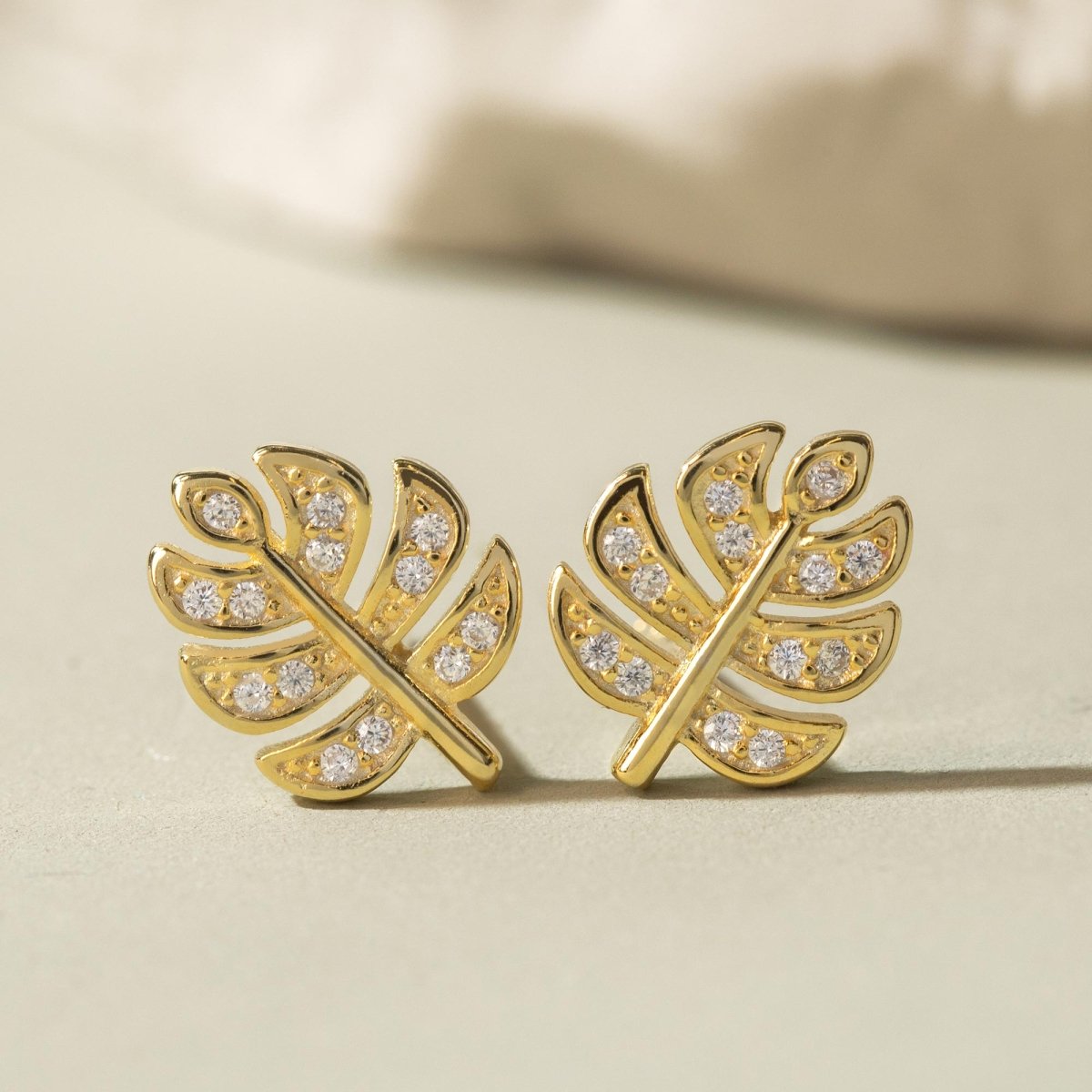 Gold vermeil monstera leaf stud earrings with clear pave stones, on a cream surface