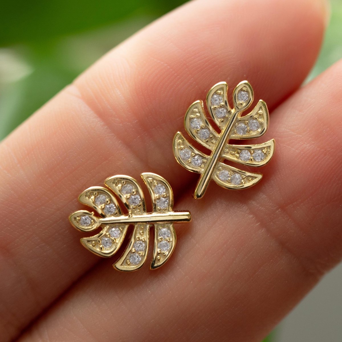 Gold vermeil monstera leaf stud earrings with clear pave stones, held on fingertips