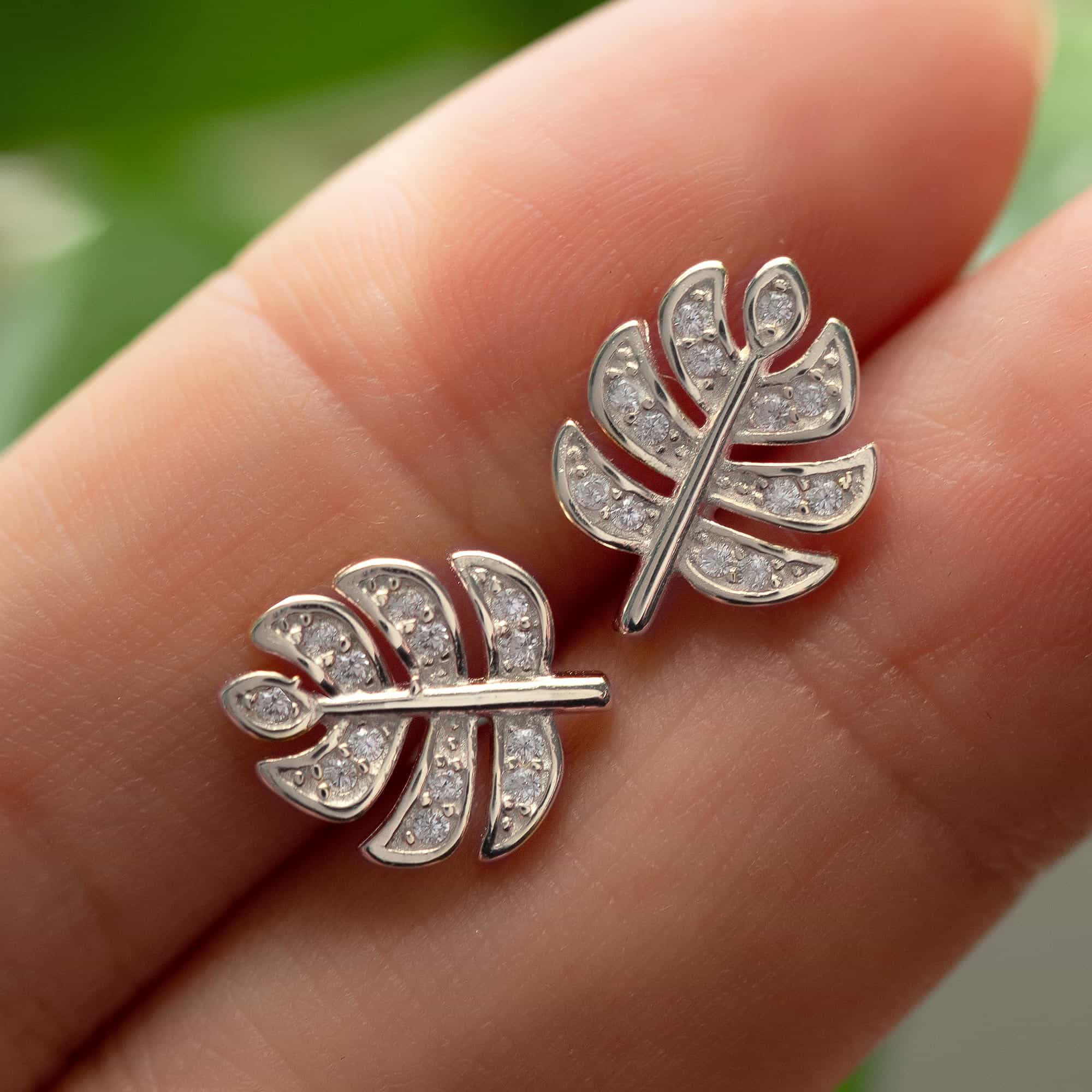 Sterling silver monstera leaf stud earrings with clear pave stones, held on fingertips