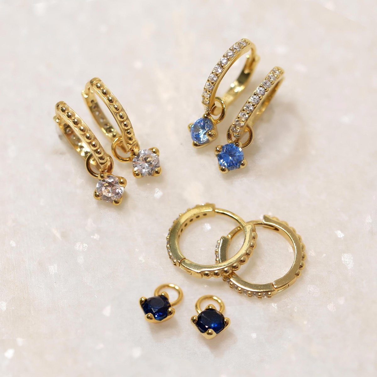 Gold vermeil pavé and beaded huggie hoops with clear and blue dangling charms, on a cream background