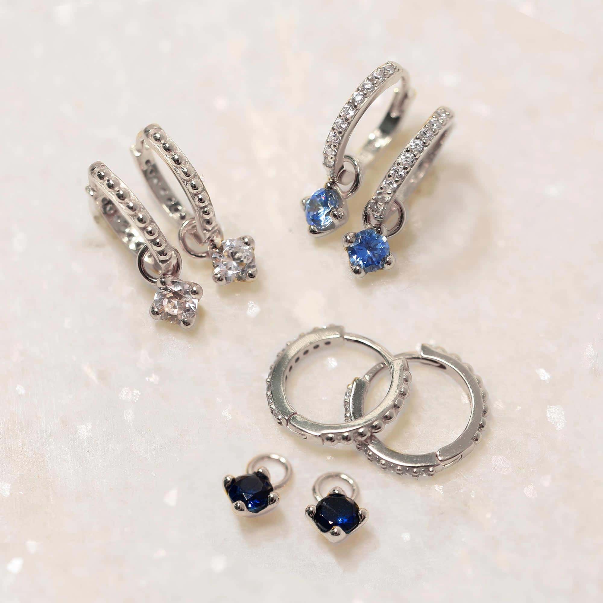 Sterling silver pavé huggie hoops with white zirconia and blue stone charms, laid on white marble