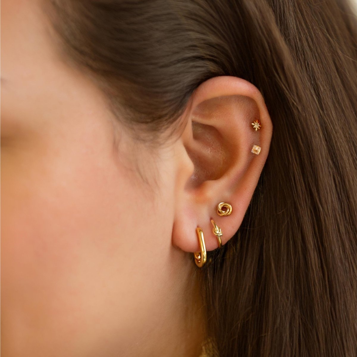14K gold U-shaped huggie hoop shown on a model's lobe with stud and knot piercings