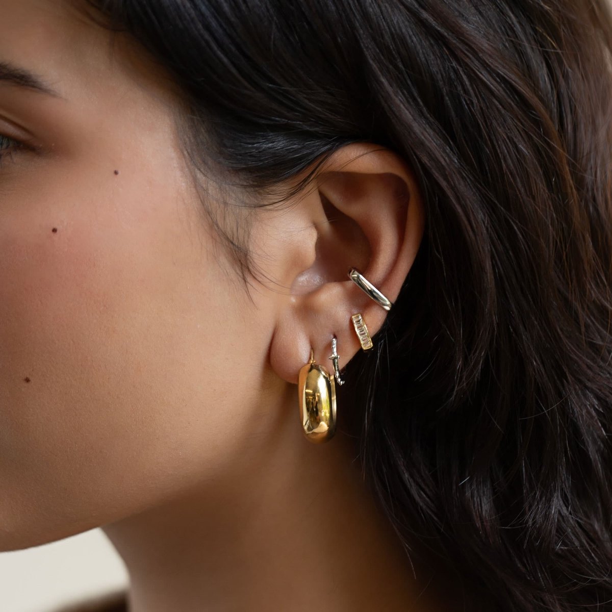 Sterling silver beaded cross huggie hoops shown in a mixed-metal ear stack on a model