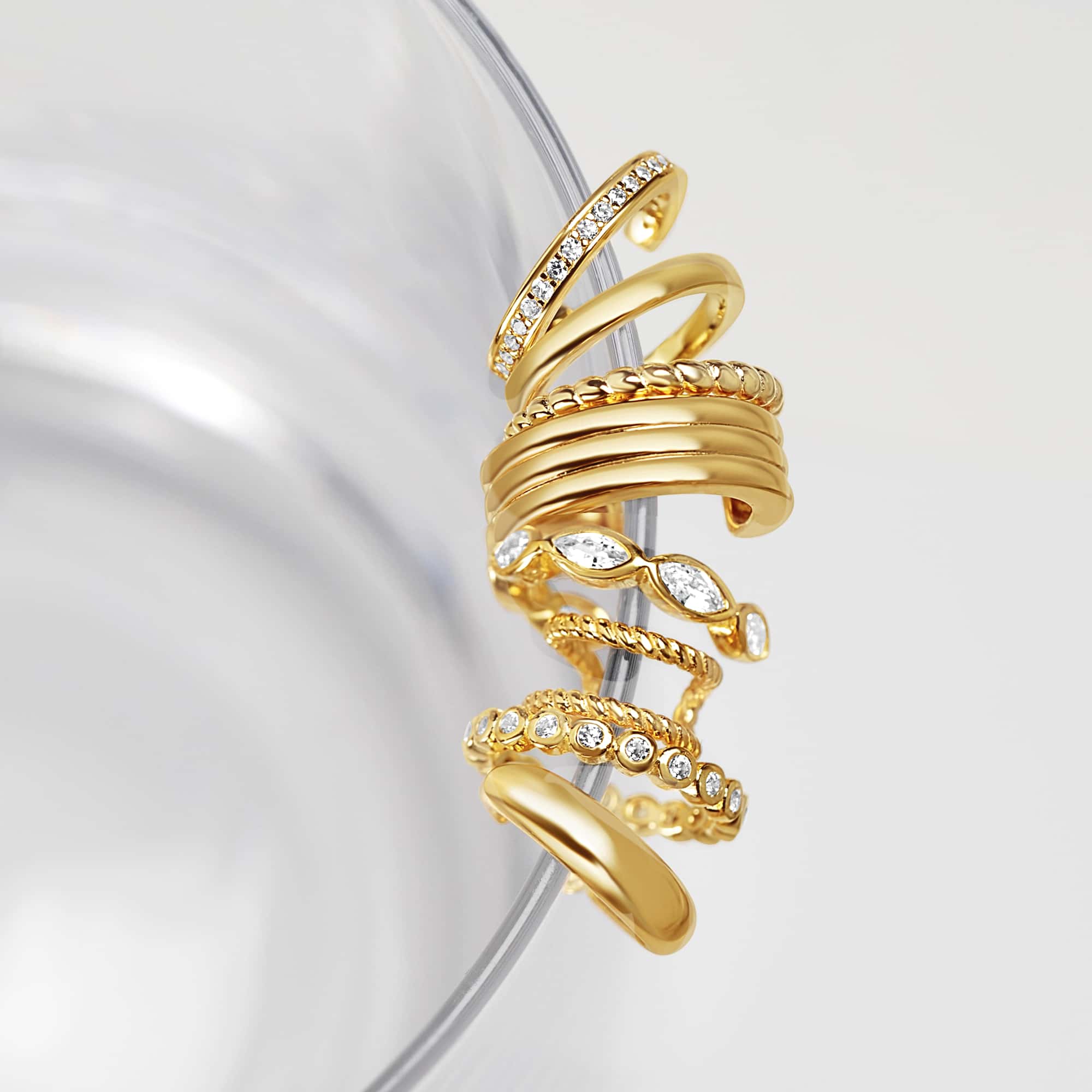 Assortment of gold ear cuffs with clear stones, rope and plain styles, coiled on a clear glass rim