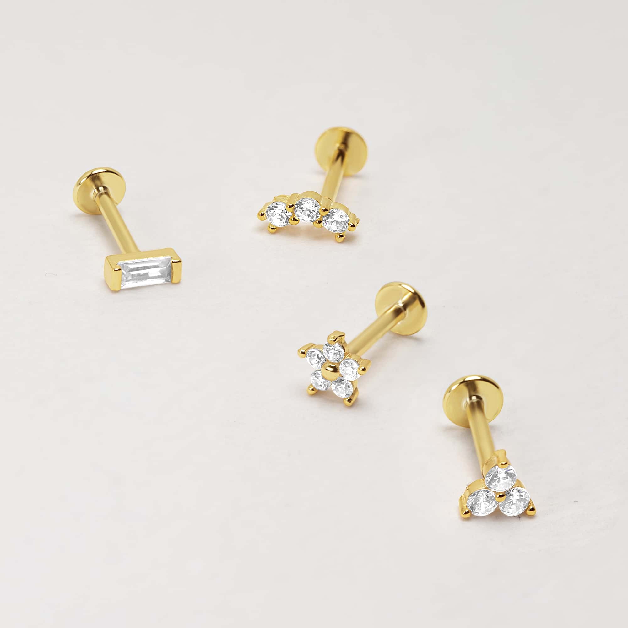 Four gold flat back studs in baguette, arc, flower and trio styles with clear stones, on a cream background