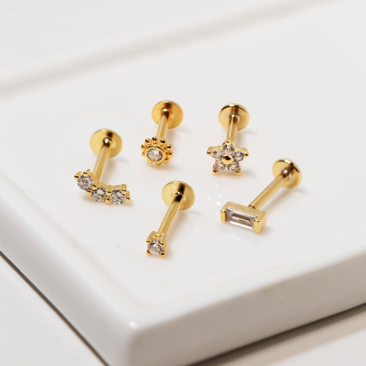 Five gold vermeil flat back studs with clear white zirconia in flower, star and bar shapes, on a cream background