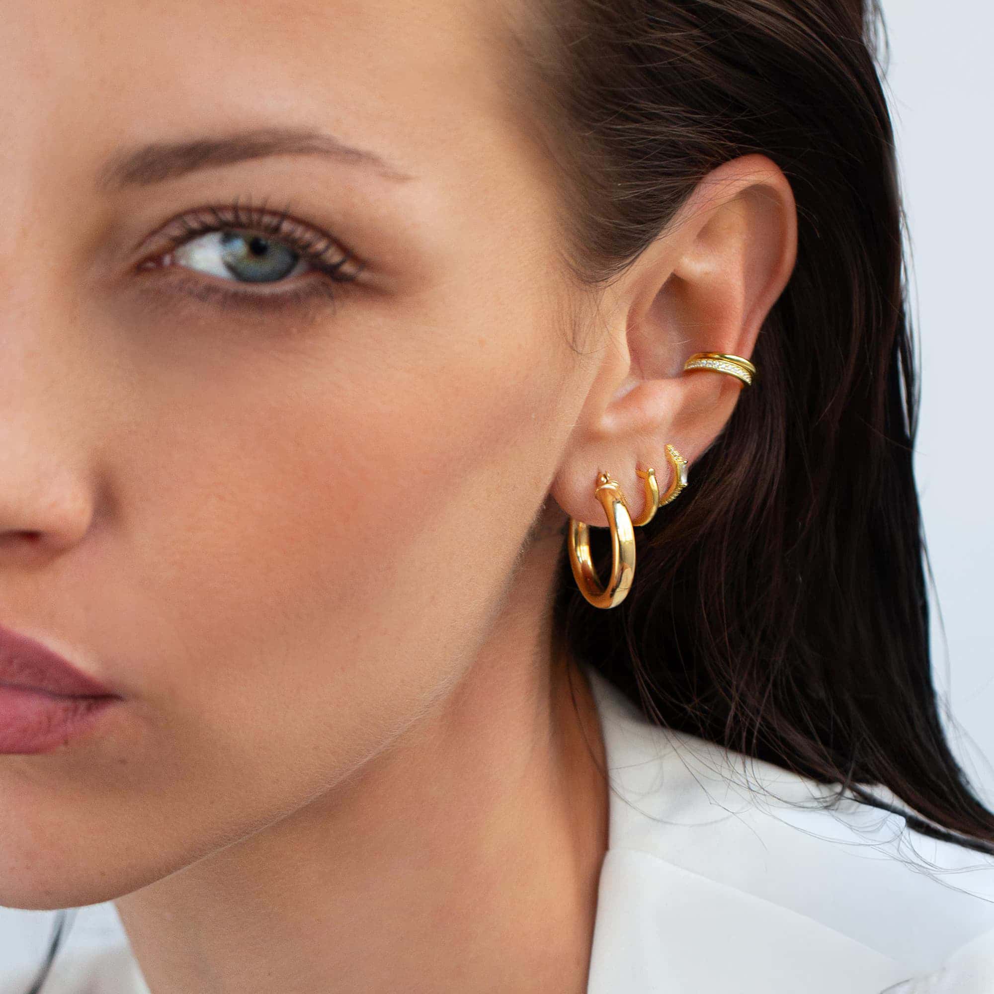 Model wearing midi thick gold hoop earrings, gold classic huggie earrings, gold gemstone baguette huggie earrings, and gold eternity and pavé ear cuff set 