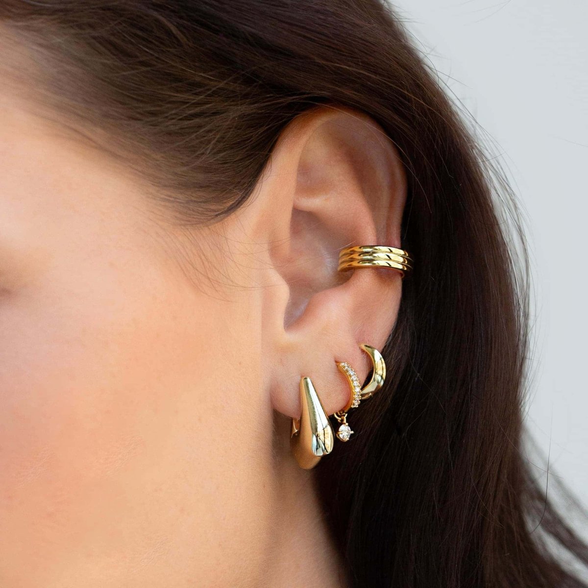 Close-up of woman's ear featuring 14K Gold Tiny Gemstone Pavé Huggies Moissanite, Delta Sculptural Small Hoops, and Gold Cuff. 