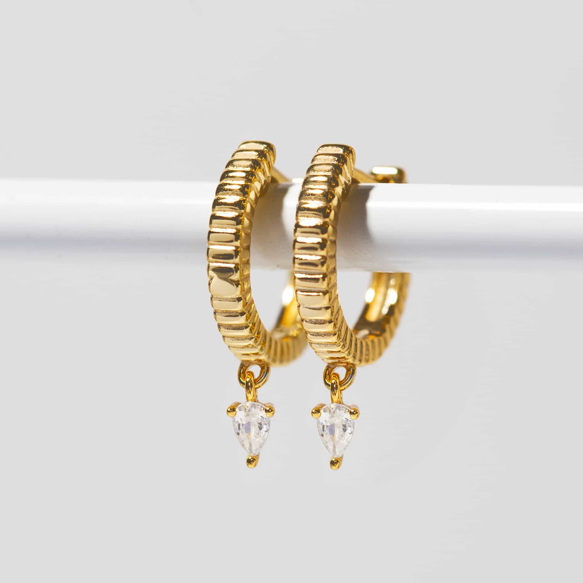 Gold vermeil ridged huggie hoops with dangling pear white zirconia, resting on a white bar