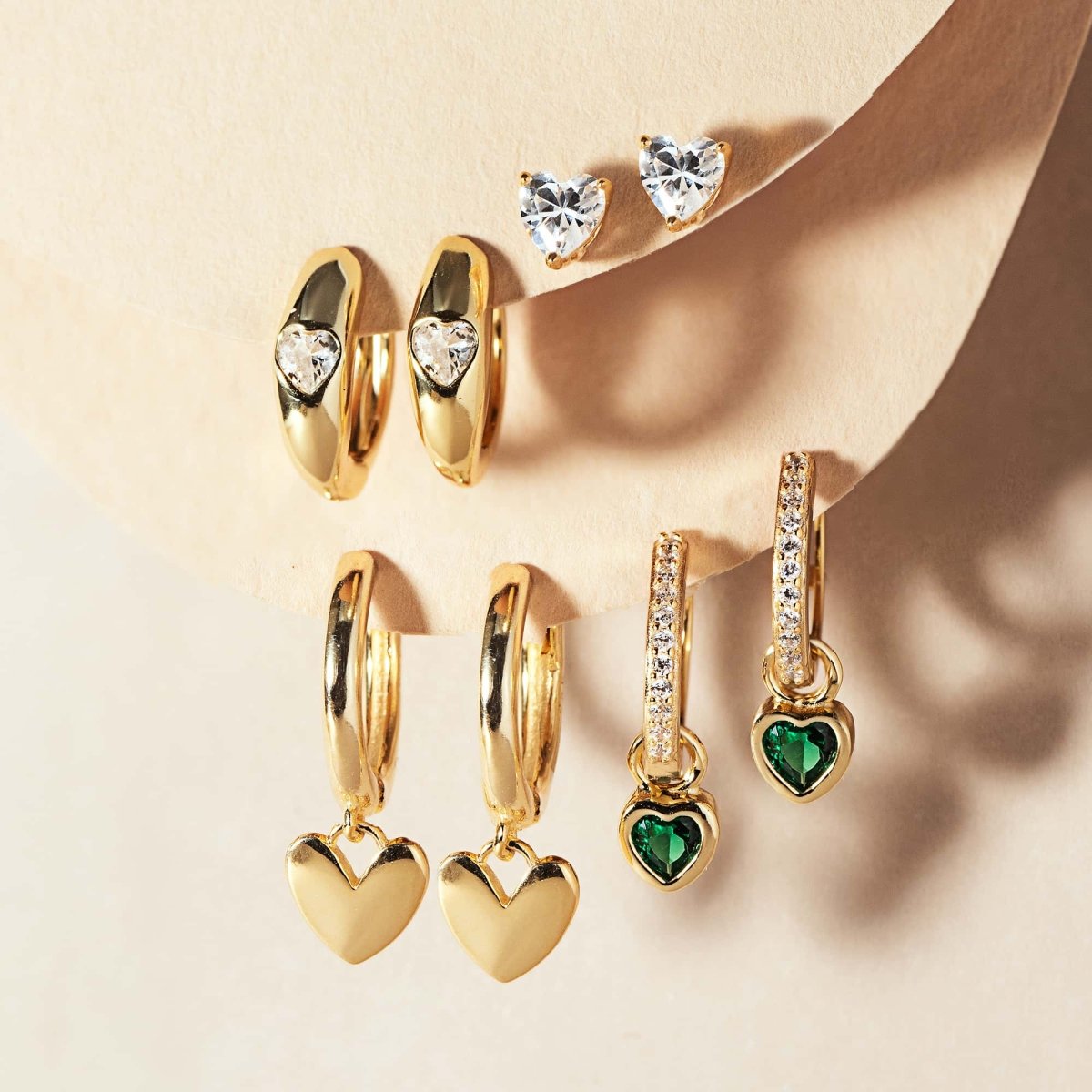 Gold vermeil heart-charm huggies shown with pavé and green stone heart styles, on a cream background