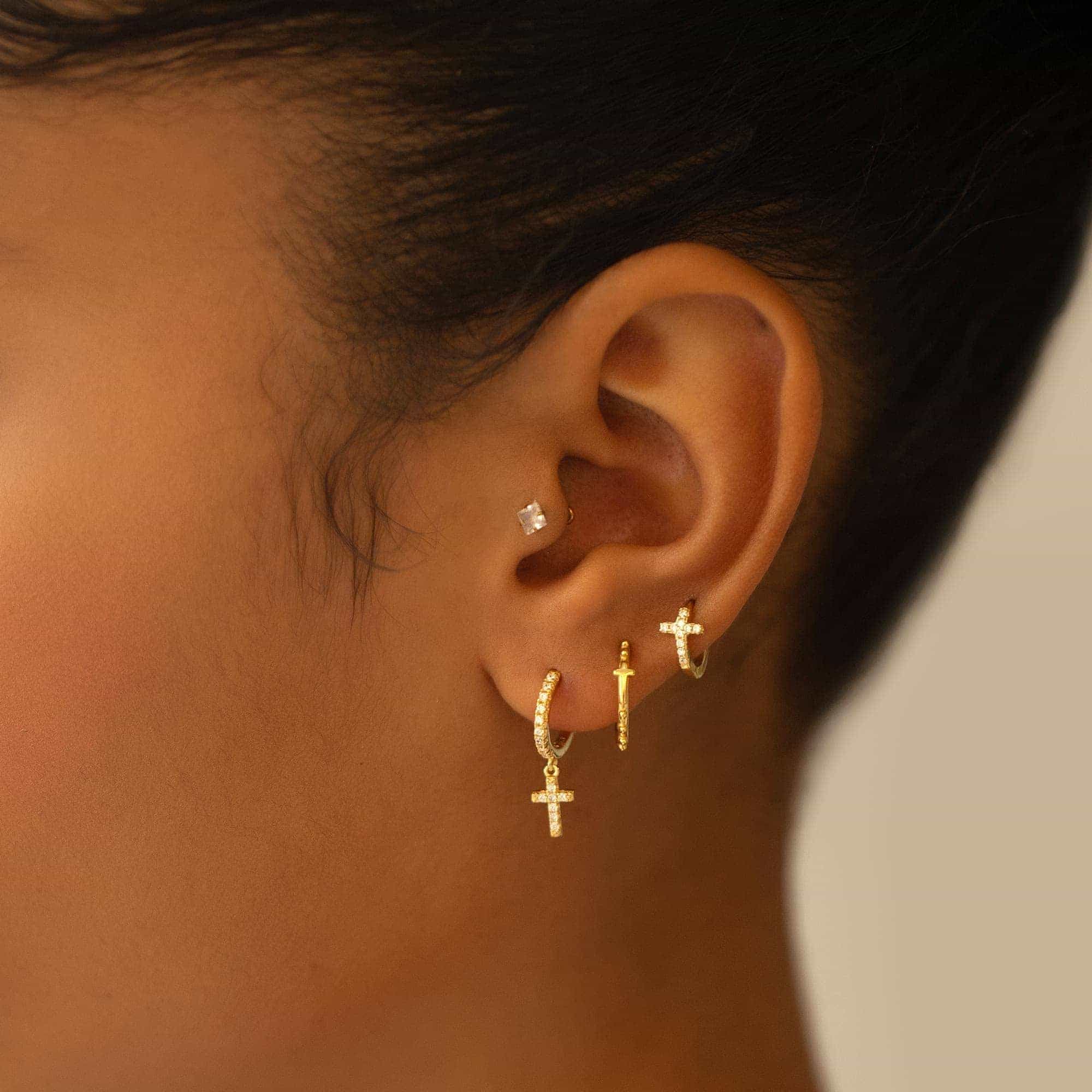 Gold cross huggie hoops, including a dangling crystal cross, stacked on a model's ear