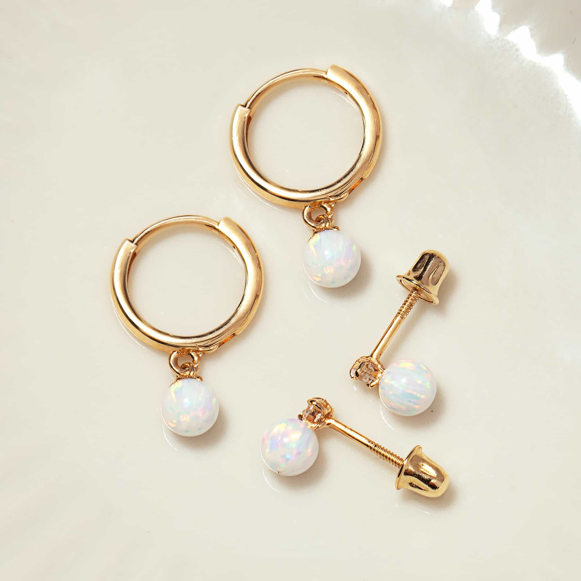 14K gold opal huggie hoops with white opal beads beside screw-back opal studs, on a cream background
