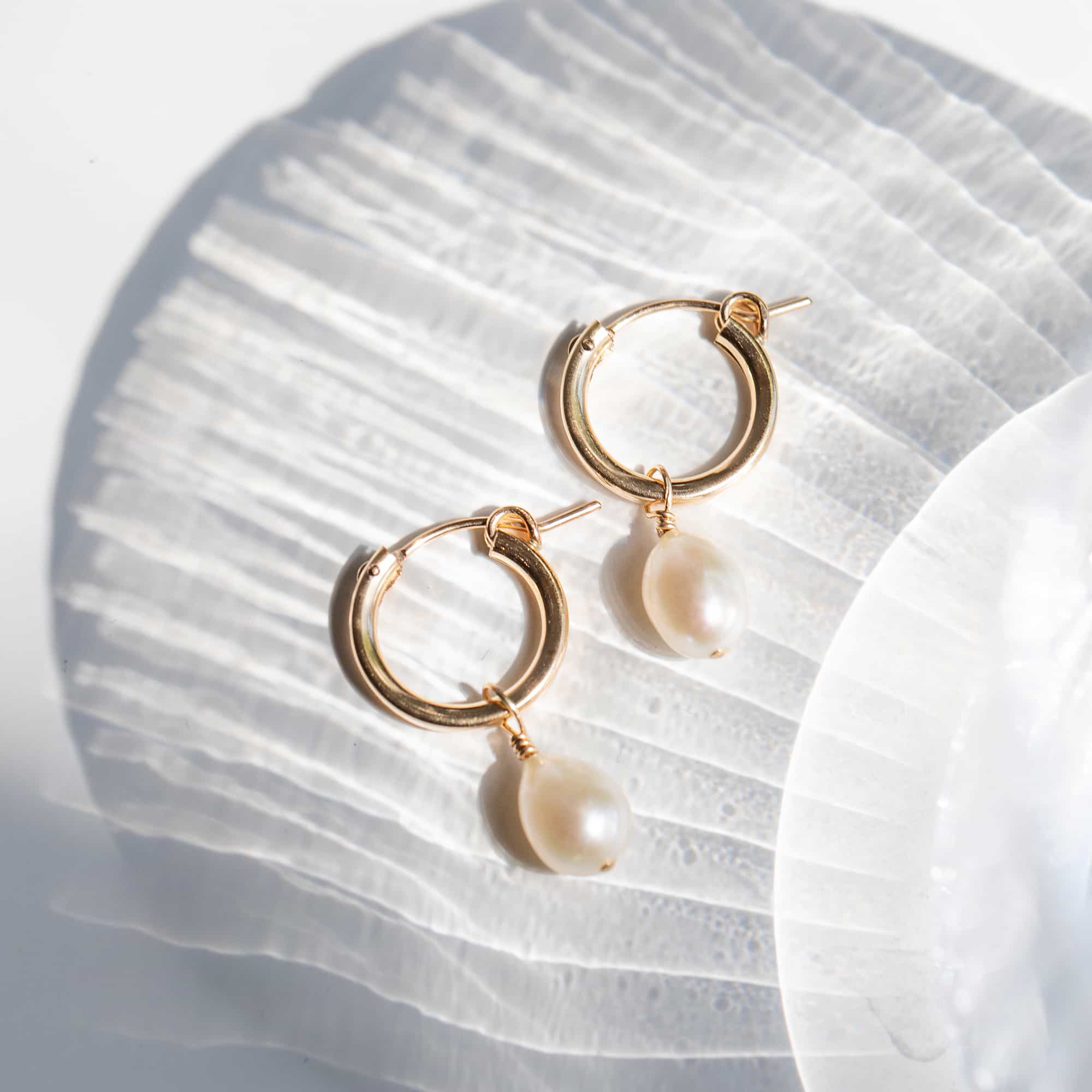 14K gold huggie hoops with a dangling white teardrop pearl, shown loose on a white surface