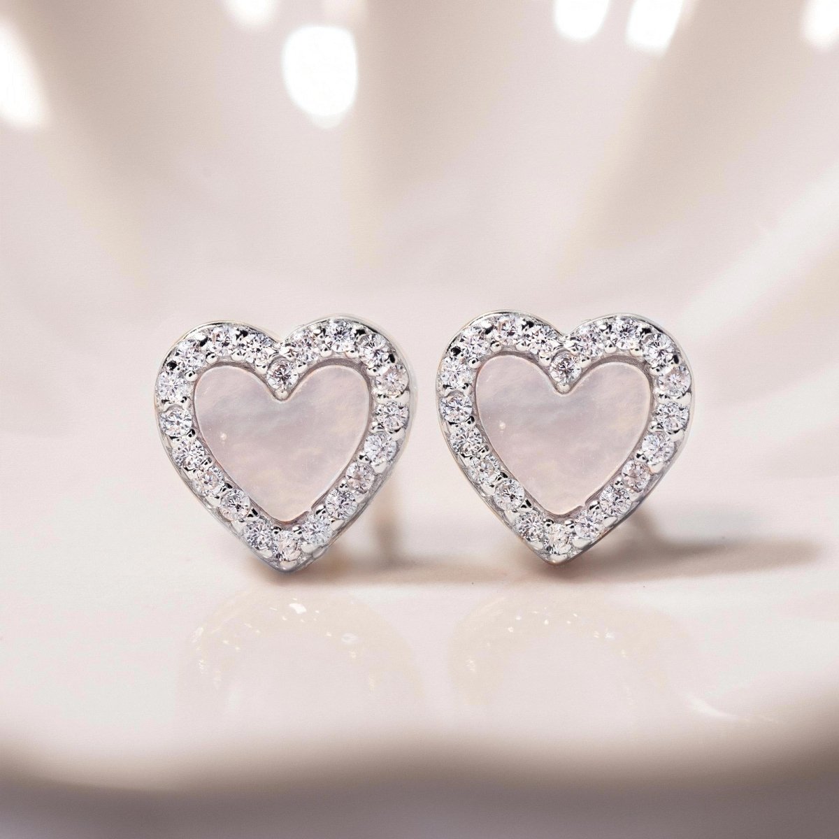 Sterling silver mother of pearl heart studs with a clear stone halo, close-up detail on a cream background