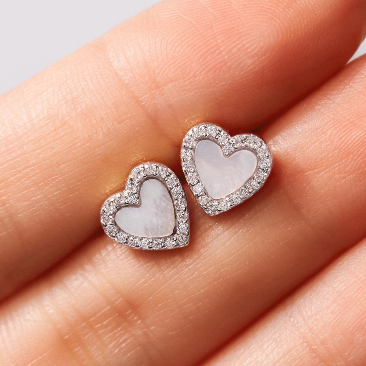 Sterling silver mother of pearl heart studs with a clear stone halo, held on fingertips